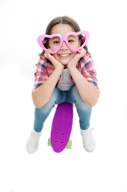 Child in heart shaped eyeglasses likes skateboarding. Joyful and happy. Kid girl casual style with penny board enjoy childhood. Girl joyful happy with skateboard isolated white. Hobby leisure concept.
