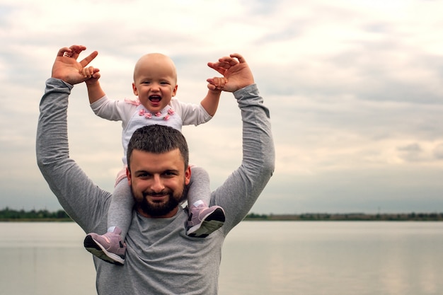 Foto un bambino sul collo di suo padre. cammina vicino all'acqua. baby e papà contro il cielo.