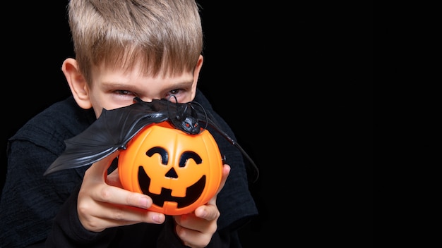 A child holding an orange pumpkin-shaped basket with a grinning face, Jack's lantern and a bat on a black background. The boy is waiting for Halloween candy. Trick or treat tradition.
