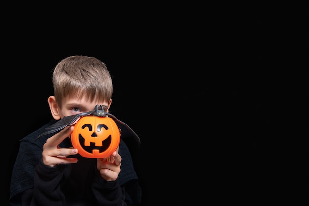 Photo a child holding an orange pumpkin-shaped basket with a grinning face, jack's lantern and a bat on a black background. the boy is waiting for halloween candy. trick or treat tradition.