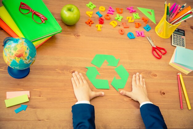 Foto bambino che tiene in mano il simbolo del riciclo oggetti scolastici sulla scrivania di legno in classe concetto educativo