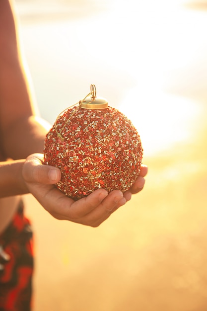 Child holding red Christmas ball in hands at the beach at sunset in California