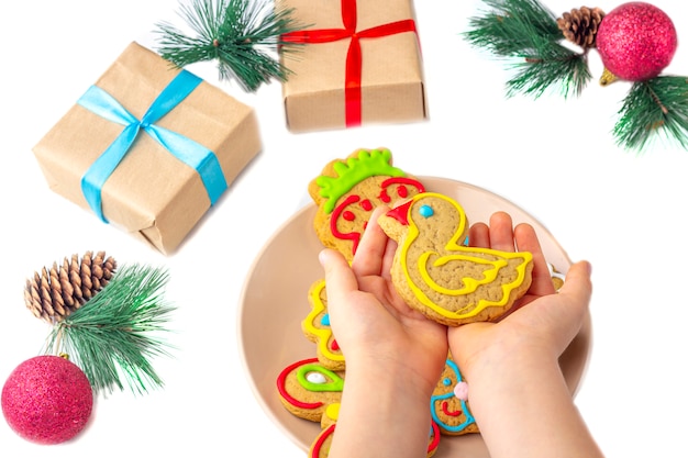 The child holds a gingerbread (cookie) on a white wooden background among fir branches and gifts. Christmas and New Year sweet gift concept. Funny sweet food close-up.