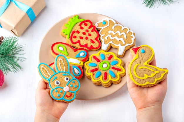 The child holds a gingerbread (cookie) on a white wooden background among fir branches and gifts. Christmas and New Year sweet gift concept.