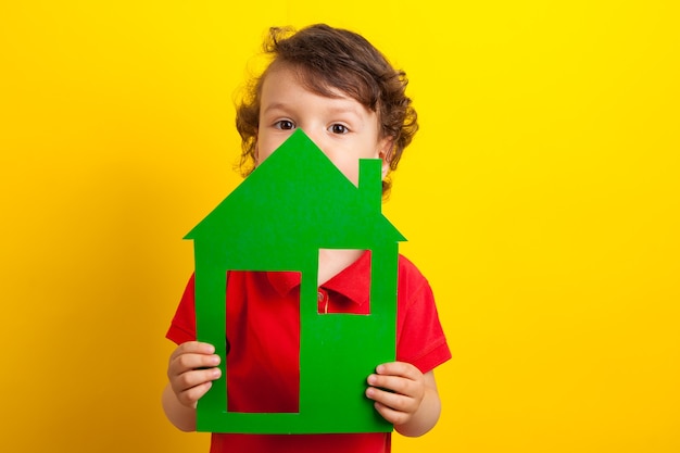 The child holds the green house on a yellow background. Conceptual photo. The boy is hiding behind the house.