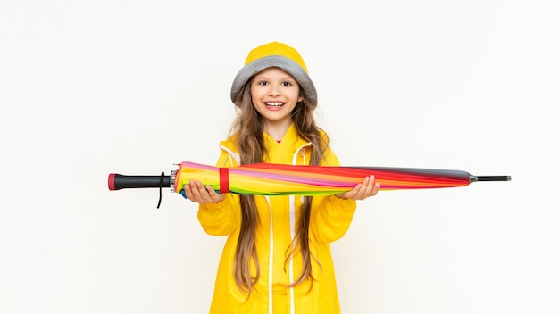 子供は傘を横に持ち、広く微笑む 孤立した白地に黄色のレインコートとパナマ帽をかぶった少女