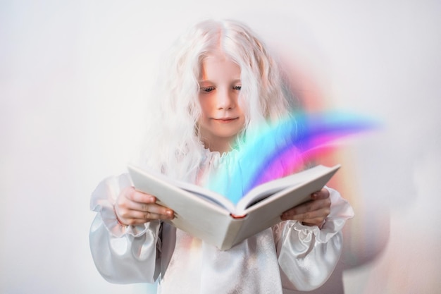 Child imagination Story time Fairytale magic Portrait of curious sweet blonde little girl in white vintage dress with bright rainbow flowing from open book on light background