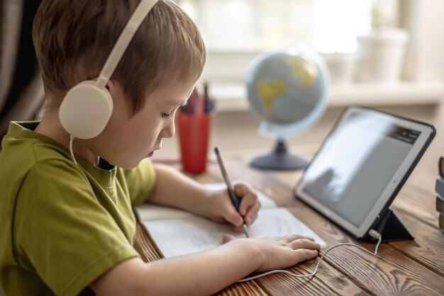 子供は緑のTシャツを着た男の子で、ヘッドホンでタブレットで宿題をしています遠隔学校教育のコンセプト