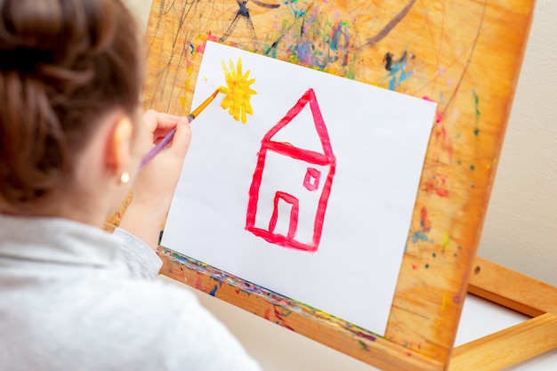 Child is drawing house with watercolors on the easel