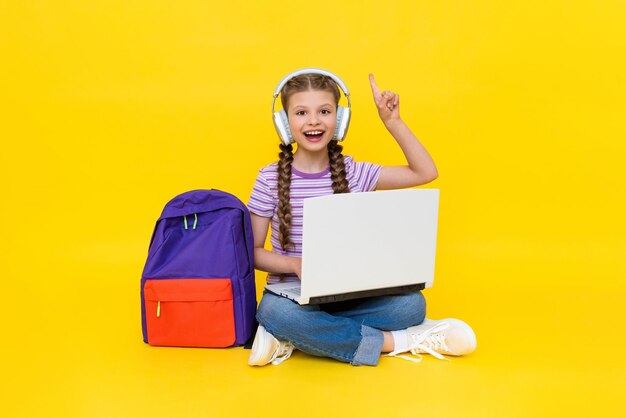 The child is engaged in an online lesson with a laptop and points up with his finger A young girl is sitting crosslegged on the floor with a backpack and headphones Yellow isolated background