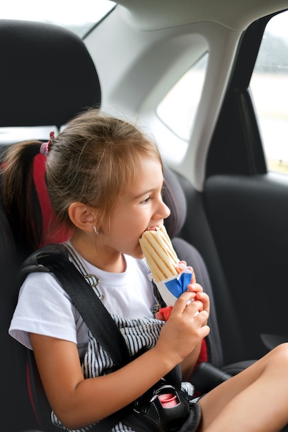 The child is fastened with a seat belt in a car seat and eats a French bun