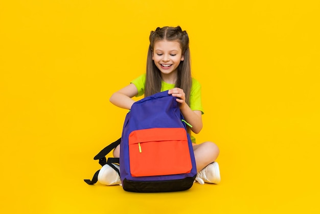 The child is going to school sitting a little girl is rummaging through a school backpack additional education for schoolchildren preparatory courses for children yellow isolated background