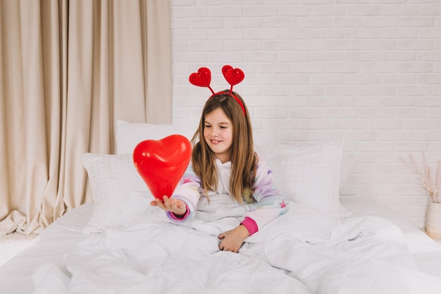 The child is holding a balloon in the shape of a heart. a little girl lies on the bed and holds a heart.Valentine's day