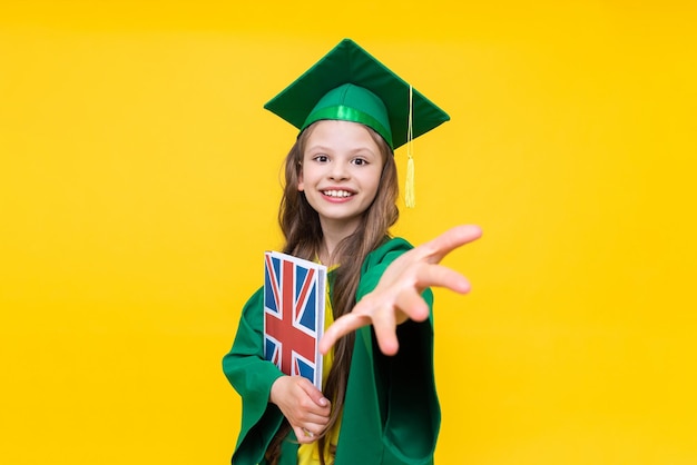 A child is holding a book on british english the student in the master's robe and hat smiles happily broadly and holds out her hand forward language school for children yellow isolated background