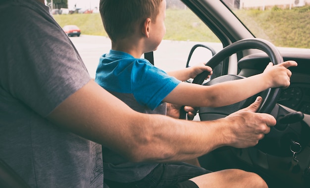 Child is sitting on the father's knees in the car and learning how to drive the car