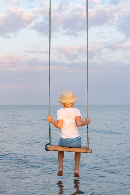 Child is sitting on rope swing over water. Sea vacation with children.
