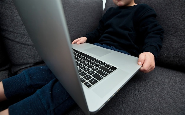 The child is sitting on the sofa holding the laptop and watches the cartoons