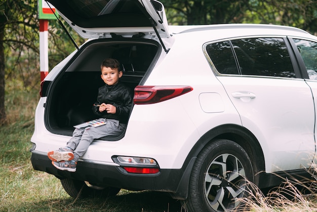 A child is sitting in the trunk of a car suv or crossover with gouache paints a car in the autumn fo...