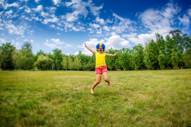 La ragazza del bambino del bambino con l'espressione e le ghirlande felici divertenti a braccia aperte della parrucca blu del pagliaccio del partito sta saltando nel parco