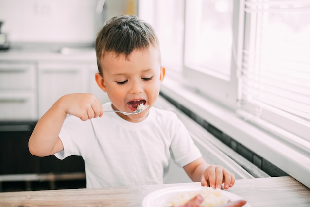 Il bambino in cucina che mangia da solo salsiccia e purè di patate