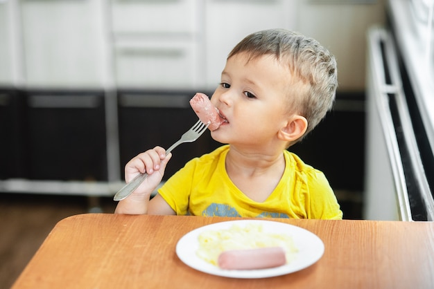 Bambino in cucina che mangia salsiccia e purè di patate