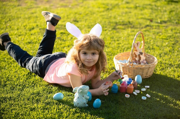 公園の草の上に横たわる子供イースターエッグ子少年バニーの耳を持つウサギの衣装で e を描く