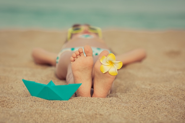 Child laying on the sand at the beach with a flower on his foot and a blue origami boat