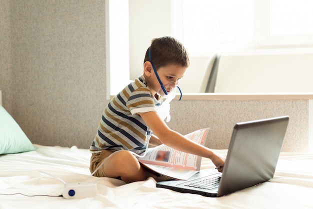 A child learns home lessons in an oxygen mask with a nibulizer, laptop and a book