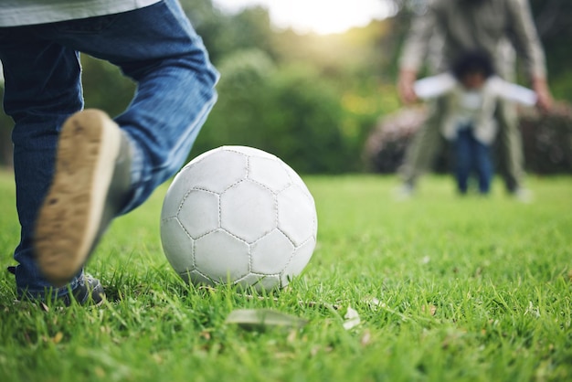 子供の足と草の上でサッカー ボールを蹴る楽しい活動の子供時代や公園で遊ぶスポーツ ゲームや屋外の自然の緑のフィールドでのスコア ポイントやゴールの試合で遊び心のある小さな男の子
