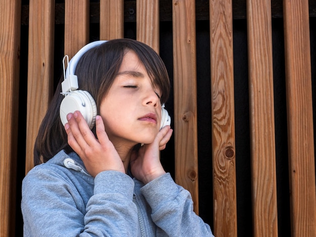 Child listening to music with white headphones in front of a wooden wall