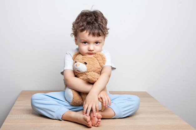 子、テーブルの上に座っている小さな男の子はクマを抱擁します。カメラを真剣に見てください。疲労と落胆。白色の背景。孤立した