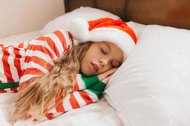 A child, a little girl in Christmas pajamas and Santa Claus hat, sleeps on the bed at home.