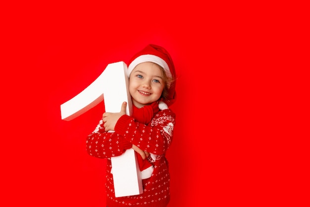 Child a little girl holding number one in Santa hat on red background, space for text