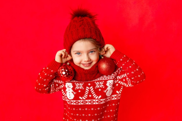 Child a little girl in winter clothes holds Christmas balls as earrings on a red background New year's concept space for text