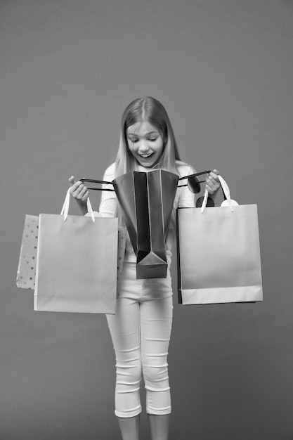 Child look in shopping bags on violet background Little shopaholic smile surprised with paper bags Childhood Shopping and sale Shopper girl with paperbags What a surprise Big sale and shopping