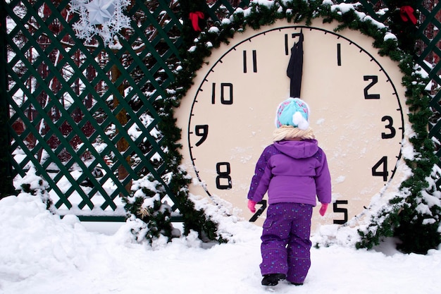 The child looks at the clock against the background of winter there is snow around time is 12