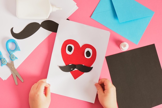 Ребенок делает открытку на день отца красное бумажное сердце с усами открытка для папы пошаговые инструкции