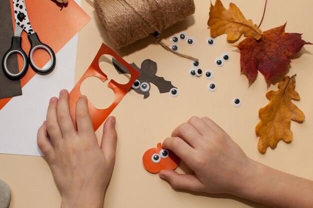 Photo a child makes a halloween craft in the form of a pumpkin garland a ghost and a bat with eyes on a beige background