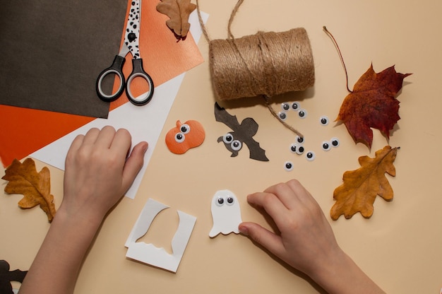 子供は、カボチャの花輪の形でハロウィーンの工芸品を作り、ベージュの背景に幽霊と目を持つコウモリを作ります