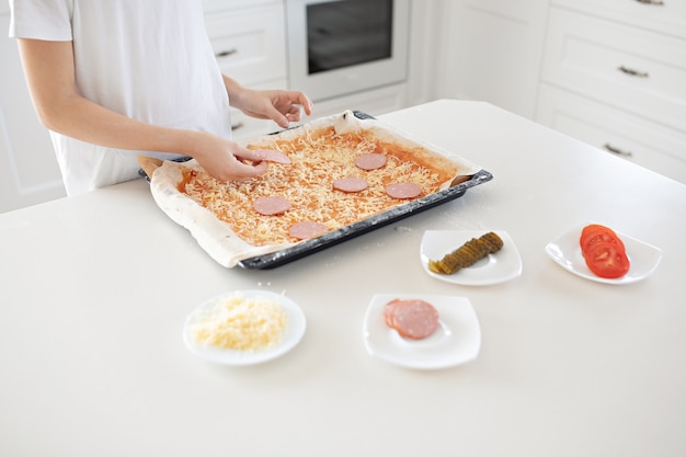 A child makes a pizza. Salami, cheese, ketchup, tomatoes, cucumbers.