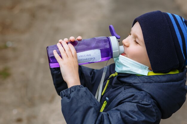 Un bambino in una maschera medica che beve acqua da una bottiglia che dice il vaccino contro il coronavirus