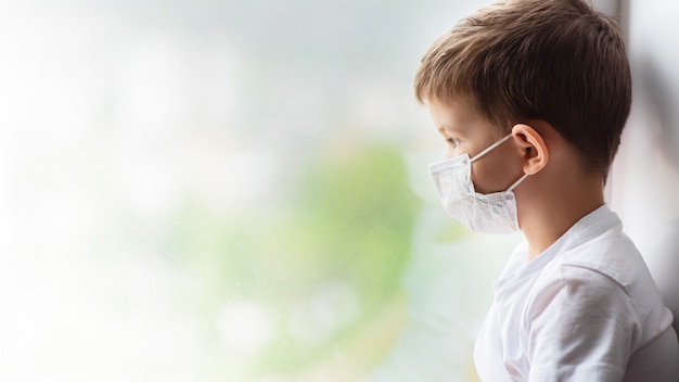 A child in a medical mask is sitting at home in quarantine because of coronavirus and covid -19 and looks out the window.