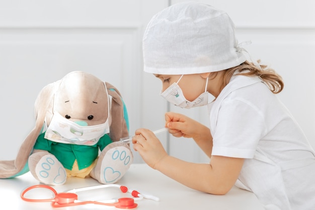 Child in medical mask playing doctor and makes injection of vaccine to toy. kid play role game at home.