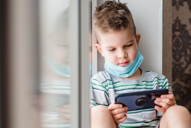 Child in a medical mask sits in quarantine and looks at the phone during the Covid-19