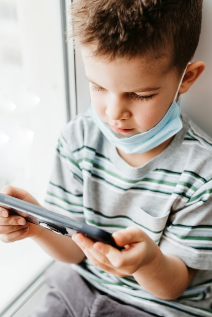 Child in a medical mask sits in quarantine and looks at the phone during the Covid-19