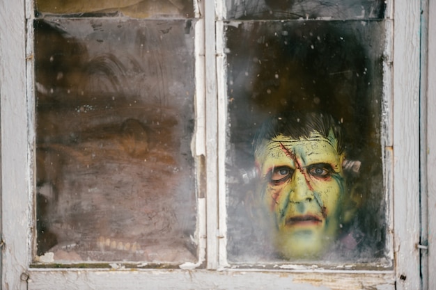 Child in a monster mask peeks out of an old dirty window in winter