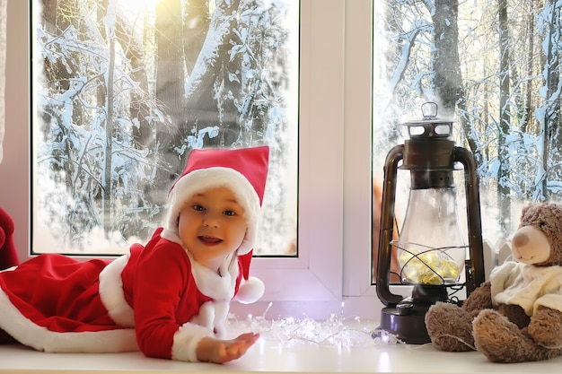 A child in the new year looks out the window. Children are waiting for Santa Claus. A child in Santa suit sits at the window.