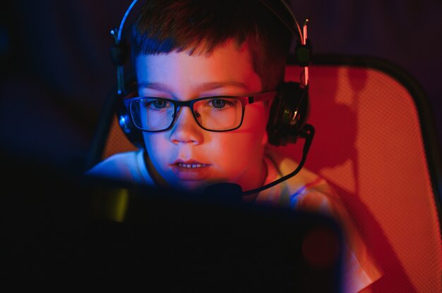 Child online broadcasts computer game, boy streams in headphones on rgb lighting background