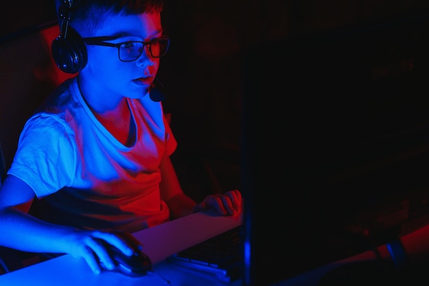 Child online broadcasts computer game, boy streams in headphones on rgb lighting background