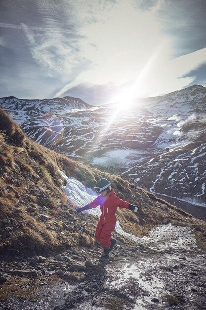 Ребенок в верхней одежде и шлеме вытягивает руки и балансирует на камнях во время прогулки возле небольшого ручья в солнечный весенний день в заснеженных горах